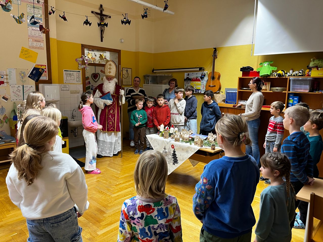 Ein Mann, der als Weihnachtsmann verkleidet ist, wird von Kindern in einem Klassenzimmer umringt, wobei ein Tisch mit Miniaturhäusern und Weihnachtsdekorationen im Vordergrund steht. Eine Frau und ein Mann stehen hinter dem Tisch, während ein Kreuz und Gitarren an den Wänden zu sehen sind.