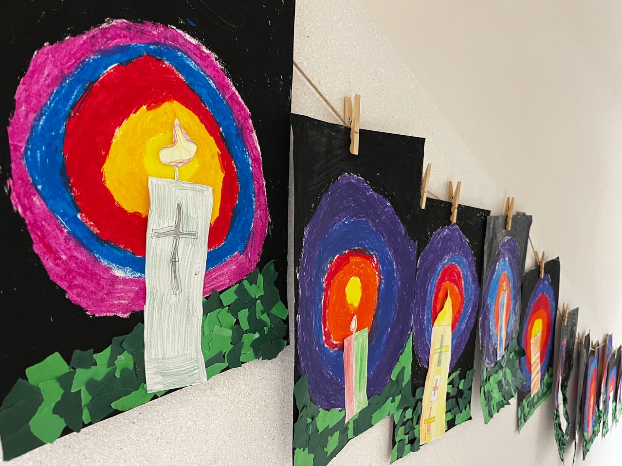 Eine Reihe von Gemälden auf schwarzem Leinwand, die Kerzen darstellen, sind an einer Wand ausgestellt. Das erste Gemälde zeigt eine Kerze, die von einem kreisförmigen Design mit einem Kreuz umgeben ist. Andere Gemälde zeigen Kerzen in verschiedenen Farben.