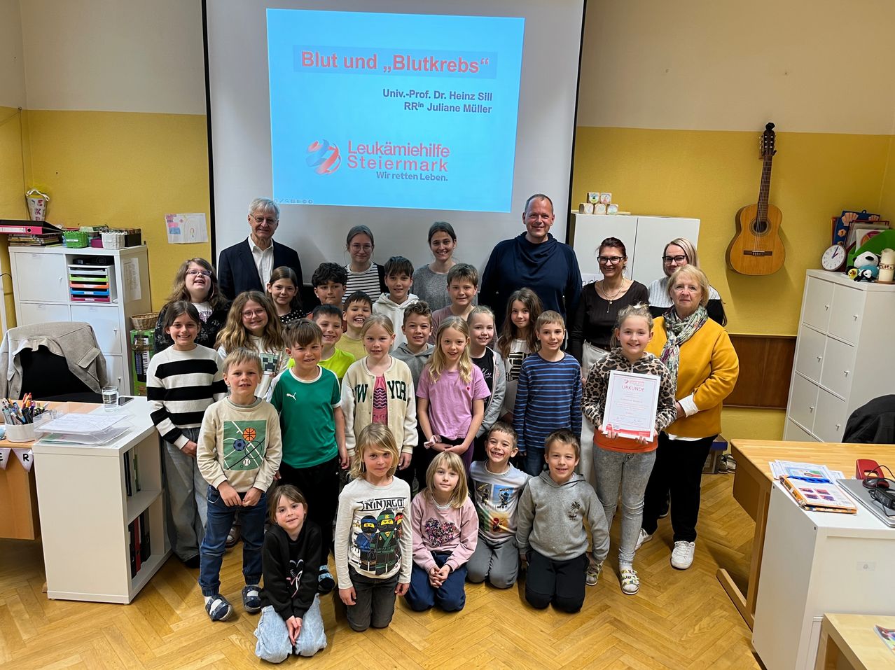 Eine Gruppe von Kindern und Erwachsenen posiert für ein Foto in einem Raum und hält ein Zertifikat. Eine Projektionsleinwand zeigt den Titel 'Blut und Leukämie'.