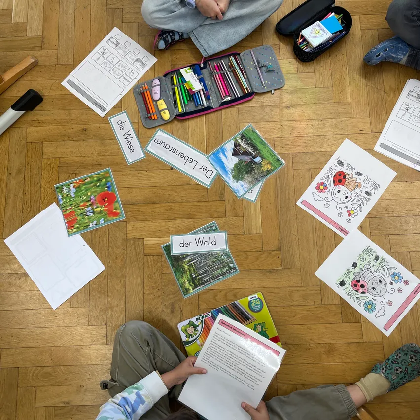 Kinder sitzen mit Malbüchern und Stiften auf dem Boden. Verschiedene Papiere und Ordner sind herum verstreut. Wörter wie 'der Wald' und 'Die Wiese' sind auf dem Boden sichtbar.
