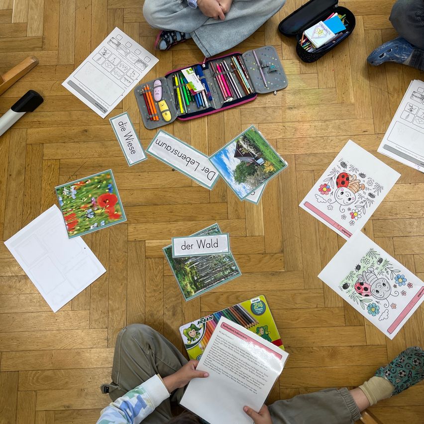 Kinder sitzen mit Malbüchern und Stiften auf dem Boden. Verschiedene Papiere und Ordner sind herum verstreut. Wörter wie 'der Wald' und 'Die Wiese' sind auf dem Boden sichtbar.