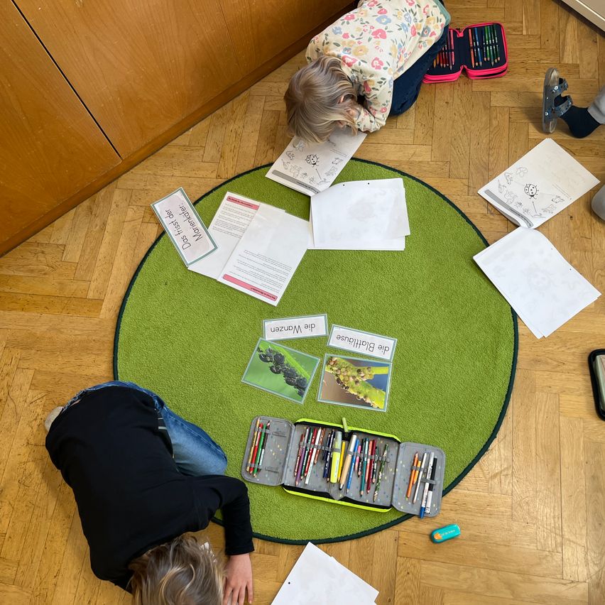 Zwei Kinder sitzen auf dem Boden und machen Kunst und Handwerk. Sie sind von Papieren, Stiften und einer Aufbewahrungsbox umgeben. Ein runder grüner Teppich liegt auf dem Boden.