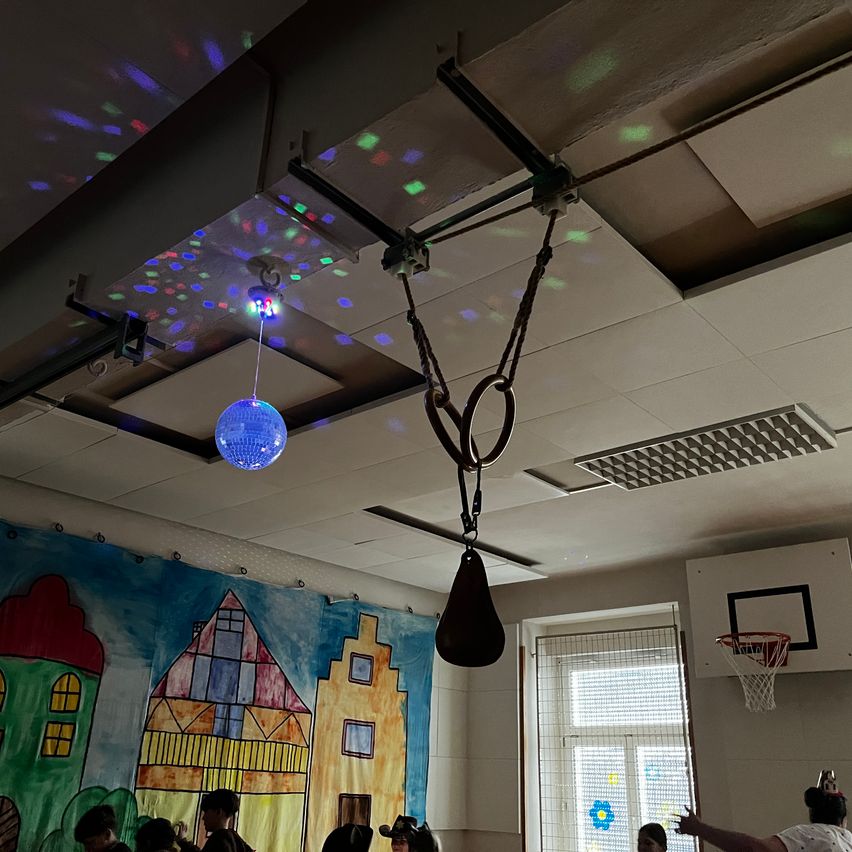 Ein Innenbereich mit einer Discokugel und farbenfrohen Lichtern, die von der Decke hängen. An der Wand befindet sich ein Wandbild von Gebäuden. Menschen sind anwesend, einige tragen Hüte. Ein Basketballkorb ist durch ein Fenster sichtbar.