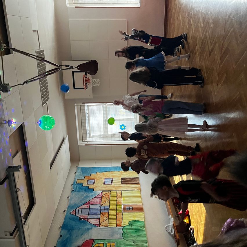 Eine Draufsicht auf einen Raum mit mehreren Menschen, die sich auf einem Holzboden versammelt haben, einer hält einen Basketballkorb, bunte Luftballons und eine Wandmalerei an der Wand.