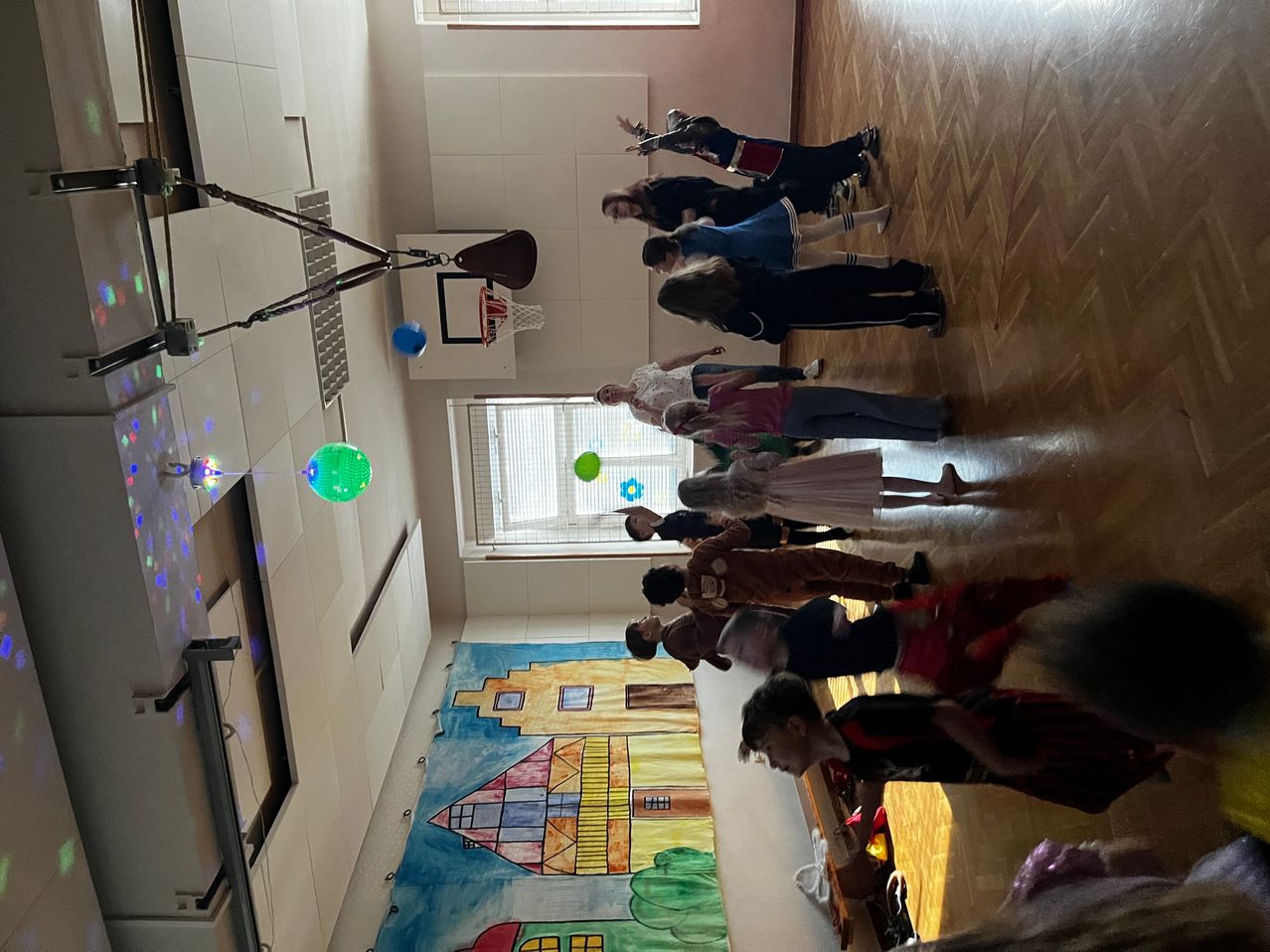 Eine Draufsicht auf einen Raum mit mehreren Menschen, die sich auf einem Holzboden versammelt haben, einer hält einen Basketballkorb, bunte Luftballons und eine Wandmalerei an der Wand.