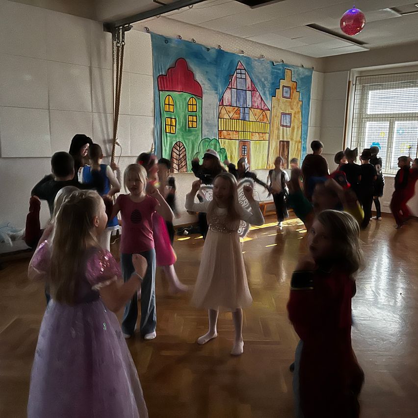 Eine Gruppe von Kindern in Kostümen tanzt in einem Raum mit Holzböden, einer Wandmalerei hinter ihnen und Sonnenlicht durch die Fenster.