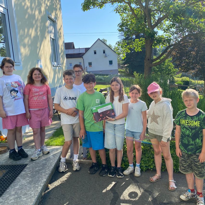 Eine Gruppe von Kindern posiert für ein Foto vor einem Haus, mit einem Garten und Bäumen im Hintergrund. Ein Kind hält ein Buch.
