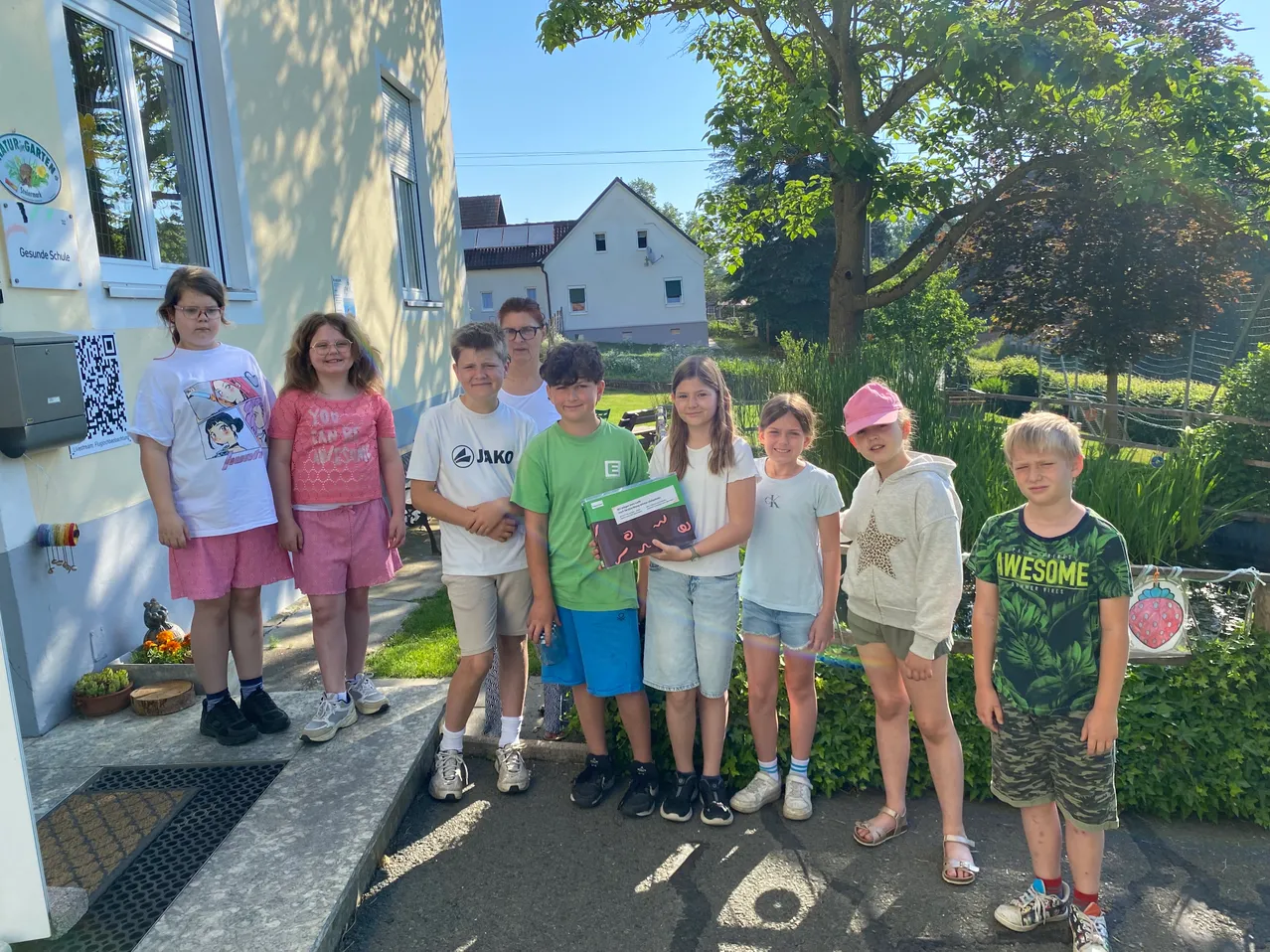 Eine Gruppe von Kindern posiert für ein Foto vor einem Haus, mit einem Garten und Bäumen im Hintergrund. Ein Kind hält ein Buch.