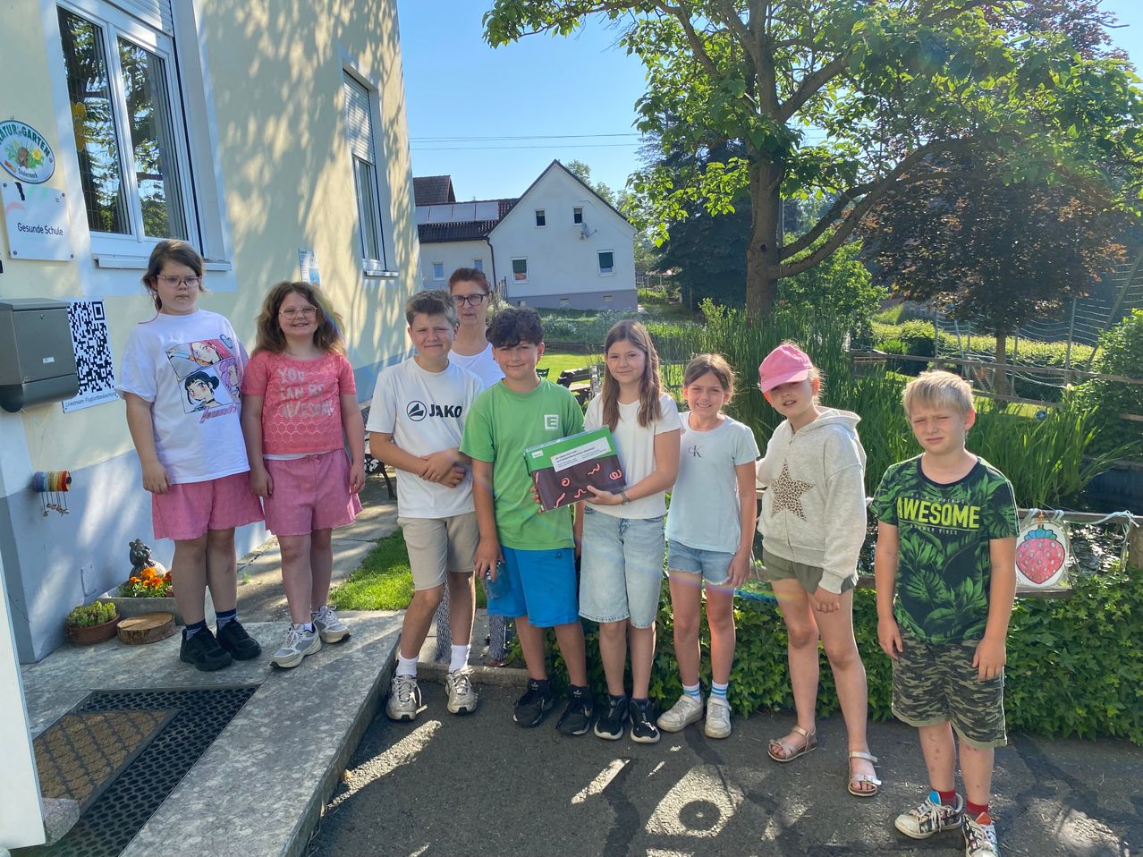 Eine Gruppe von Kindern posiert für ein Foto vor einem Haus, mit einem Garten und Bäumen im Hintergrund. Ein Kind hält ein Buch.