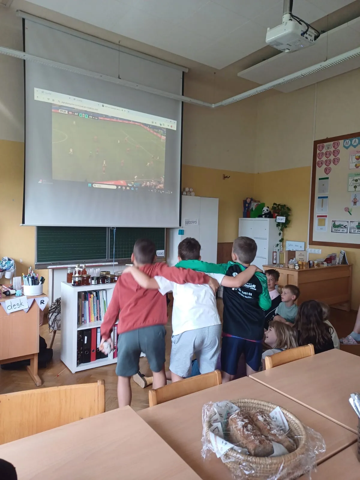 Kinder versammeln sich in einem Klassenzimmer um einen Projektor, um ein Fußballspiel zu sehen. Der Raum hat Holztische, Regale und eine Ankündigungstafel.