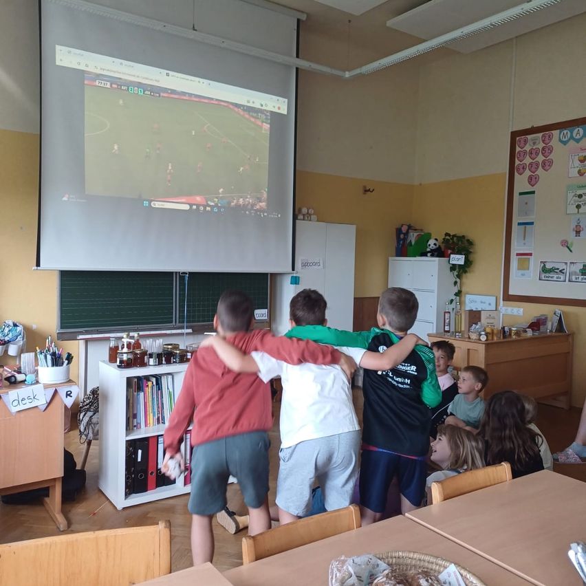 Kinder versammeln sich in einem Klassenzimmer um einen Projektor, um ein Fußballspiel zu sehen. Der Raum hat Holztische, Regale und eine Ankündigungstafel.
