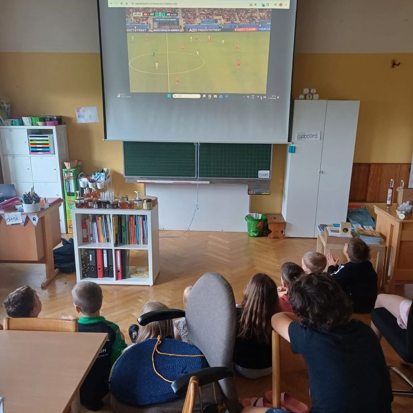 In einem Klassenzimmer sitzen Kinder auf dem Boden und schauen ein Fußballspiel auf einer Projektionsleinwand. Im Hintergrund befinden sich Regale, ein Schreibtisch und ein Schrank.