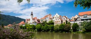 Eine Stadt mit bunten Gebäuden und einem Turm liegt am Ufer eines Flusses unter einem blauen Himmel mit verstreuten Wolken.