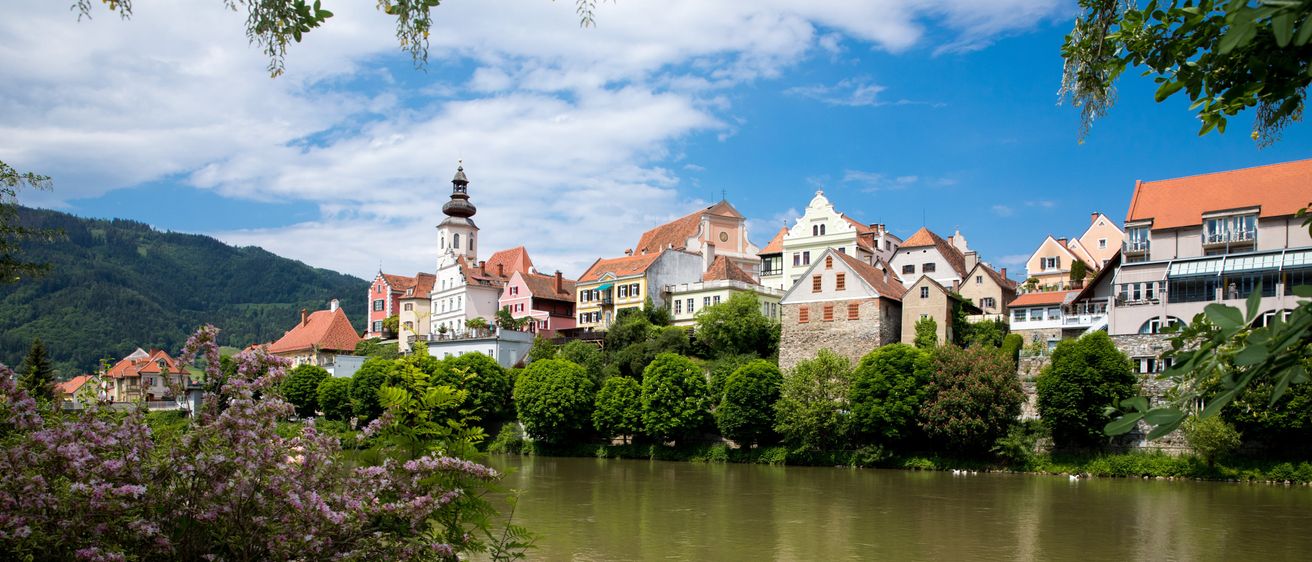 Eine Stadt mit bunten Gebäuden und einem Turm liegt am Ufer eines Flusses unter einem blauen Himmel mit verstreuten Wolken.