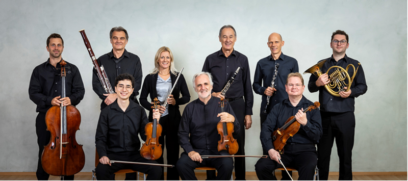 Eine Gruppe von Musikern in Schwarz posiert für ein Foto. Sie halten verschiedene Instrumente wie Cello, Geige und Horn. Die Musiker stehen und sitzen auf Stühlen vor einer weißen Wand.