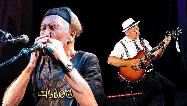 Zwei Musiker treten auf einer Bühne auf, einer spielt Gitarre und der andere Mundharmonika. Der Gitarrist trägt einen weißen Hut und ein weißes Hemd, während der Mundharmonikaspieler ein blaues Hemd trägt.