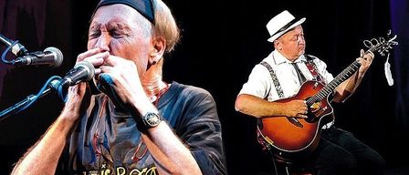 Zwei Musiker treten auf einer Bühne auf, einer spielt Gitarre und der andere Mundharmonika. Der Gitarrist trägt einen weißen Hut und ein weißes Hemd, während der Mundharmonikaspieler ein blaues Hemd trägt.