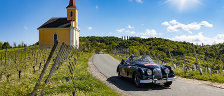 A classic car drives along a rural road with a yellow church and vineyard in the background under a bright sky.