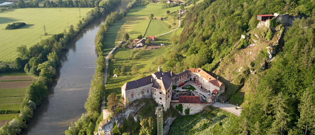 Eine Luftaufnahme einer Burg auf einem Felsen oberhalb eines Flusses, mit üppigen grünen Feldern und einer kurvenreichen Straße im Hintergrund.