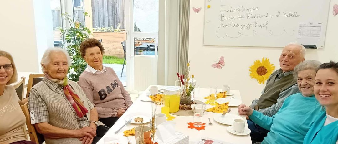 Eine Gruppe älterer Personen sitzt um einen Tisch herum und genießt ihr Frühstück. Der Tisch ist mit Tellern, Tassen und verschiedenen Speisen gedeckt. An der Wand dahinter ist eine Tafel mit handgeschriebenem Text angebracht.