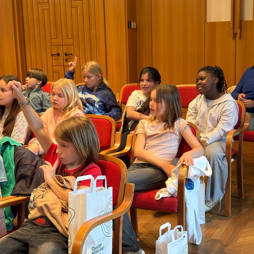 Eine Gruppe von Kindern sitzt in Stühlen, möglicherweise in einem Klassenzimmer. Einige heben ihre Hände. Sie scheinen an einer Diskussion oder Aktivität teilzunehmen. Jedes Kind hält einen Papierbeutel.
