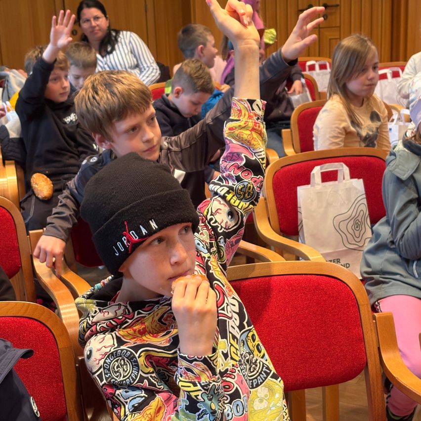 Kinder in einem Klassenzimmer sitzen auf Stühlen, einige heben die Hände, während andere Snacks essen. Ein Junge in einer Jordan-Mütze schaut sich etwas in der Hand an. Eine Frau im Hintergrund lächelt.