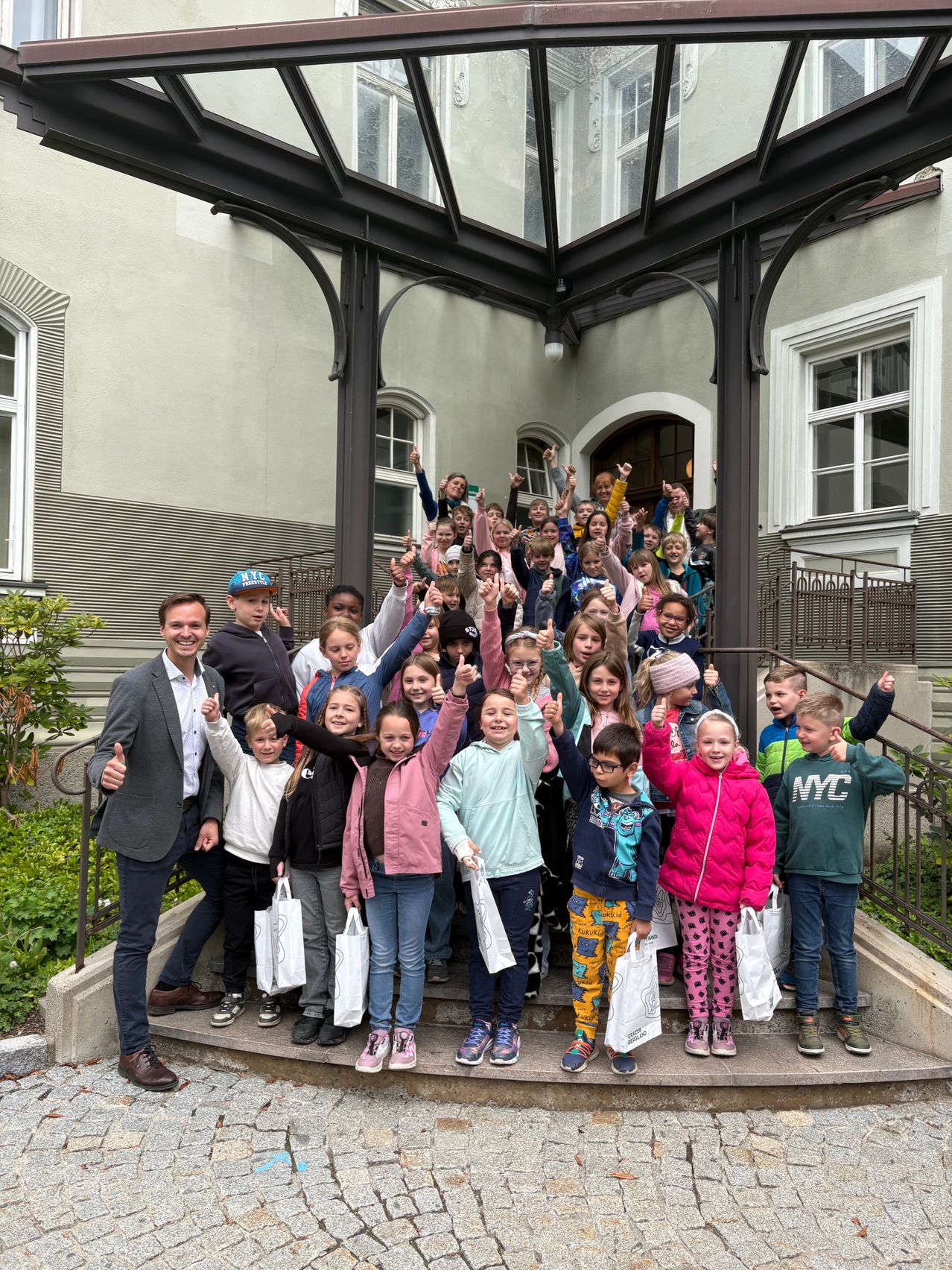 Eine große Gruppe von Kindern und Erwachsenen posiert für ein Foto auf den Stufen eines Gebäudes. Alle lächeln und heben ihre Hände. Die Erwachsenen stehen auf den unteren Stufen, während die Kinder auf den oberen Stufen stehen.