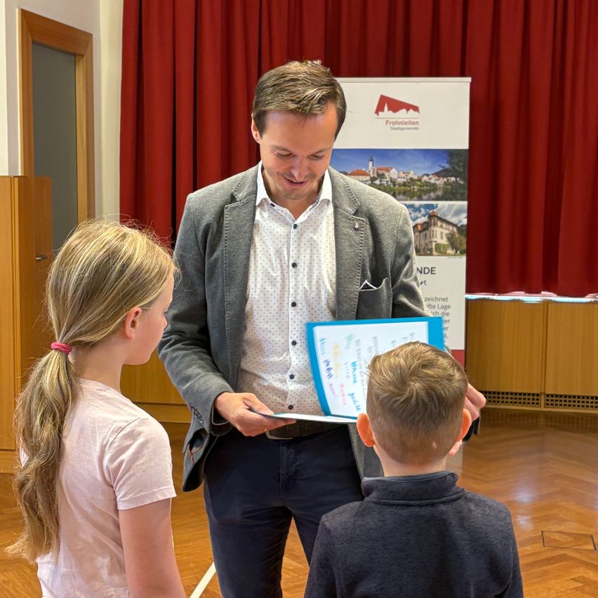 Ein Mann im Anzug steht zwischen zwei Kindern, lächelt und schaut auf ein blaues Dokument. Hinter ihm befindet sich ein Schild mit Logo und Bild. Der Raum hat einen Holzboden und rote Vorhänge.