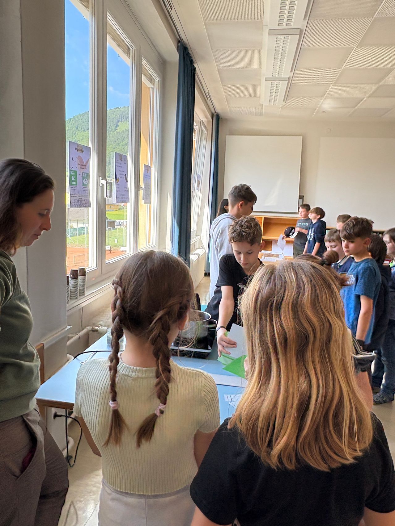 Eine Gruppe von Kindern in einem Klassenzimmer, einige versammelt um einen Tisch, andere stehen in einer Reihe. Eine Frau mit geflochtenen Haaren schaut die Kinder an. Der Raum hat Fenster mit Blick auf ein Feld und Berge.