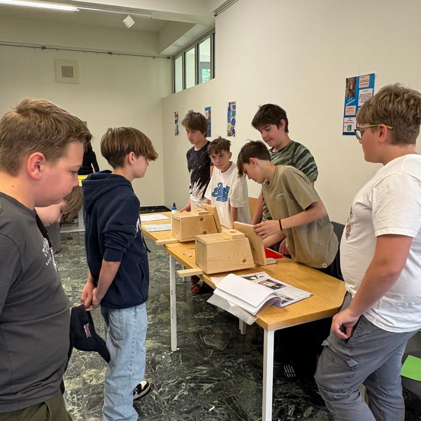 Eine Gruppe junger Jungen ist um einen Tisch versammelt und untersucht kleine Holzkisten, während eine Person mit Brille in der Nähe steht.