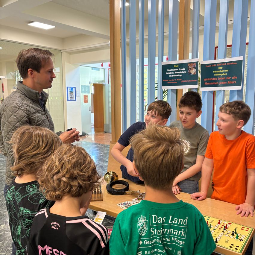 Ein Mann erklärt etwas einer Gruppe von Kindern um einen Tisch mit einem Spiel herum. Die Kinder hören aufmerksam zu. An der Wand dahinter befinden sich Poster.