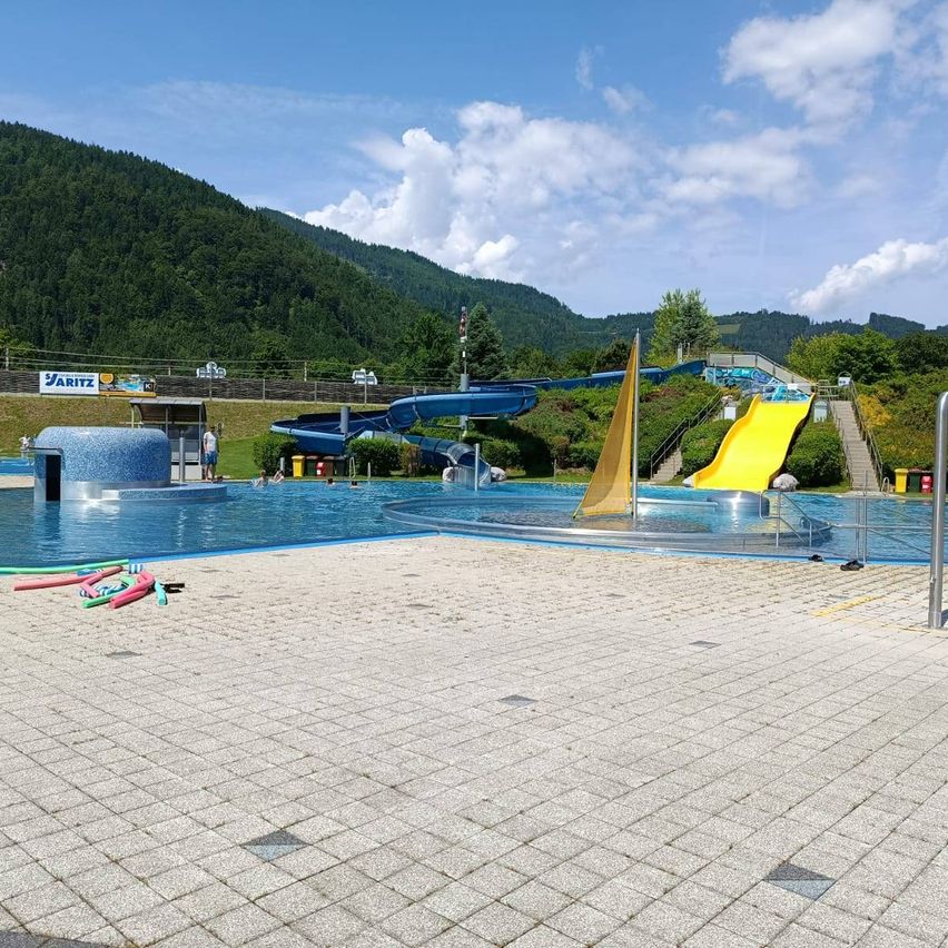 Ein Freibad mit einer blauen Rutsche und einer gelben Rutsche. Der Pool ist von einem Ziegelweg umgeben und hat einen Fahnenmast. In der Ferne befindet sich ein Berg mit Bäumen und Pflanzen.