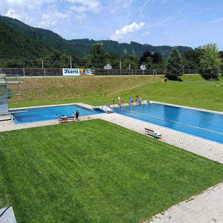 Luftaufnahme eines Freibades mit Sprungturm, umgeben von saftigem grünem Rasen, einer Bank und mehreren Schwimmern. Im Hintergrund befinden sich Berge.
