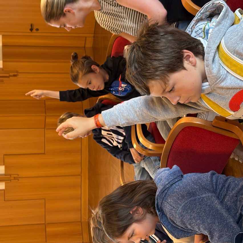 Mehrere Kinder sitzen auf Stühlen, wobei ein Junge ein Objekt genau betrachtet. In der Nähe zeigt ein Mädchen auf etwas, während eine Frau nach vorne gelehnt ist und sich im Gespräch befindet. Der Raum ist mit Holzwänden und Stühlen ausgestattet.