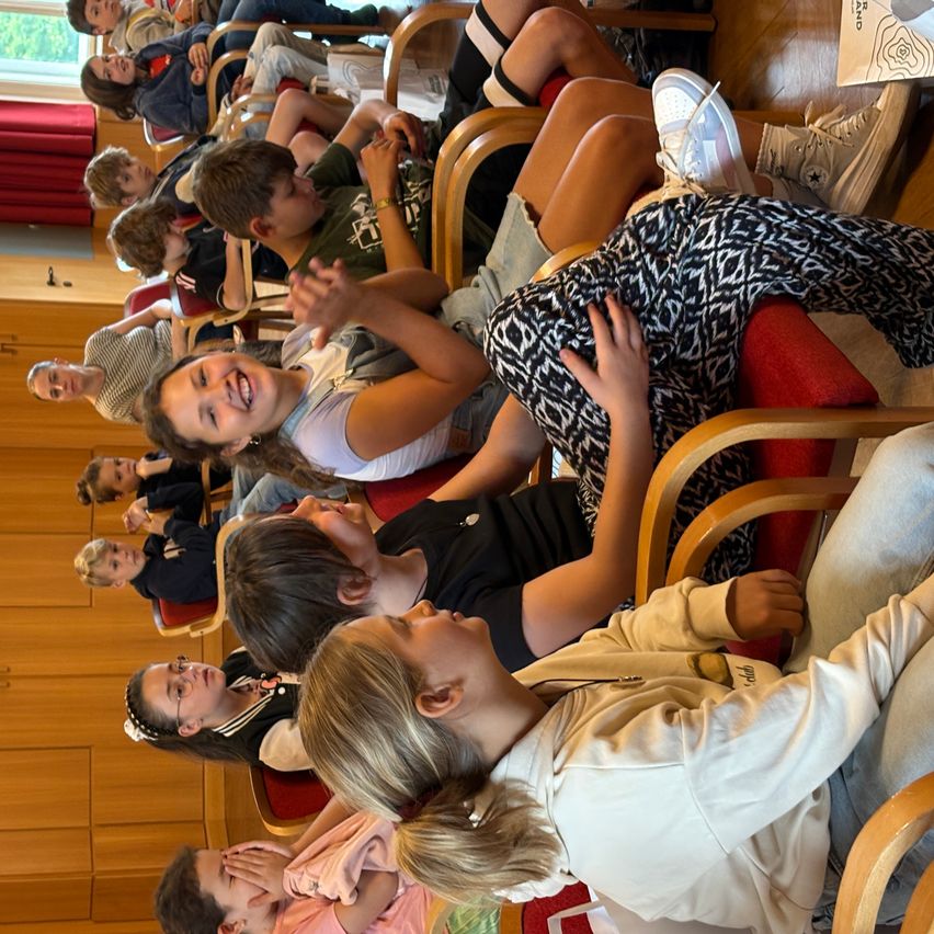 Mehrere Kinder sitzen in Stühlen, einige lächeln und schauen sich etwas an. Ein Junge vorne trägt eine Brille und ein Mädchen neben ihm hebt die Hand.