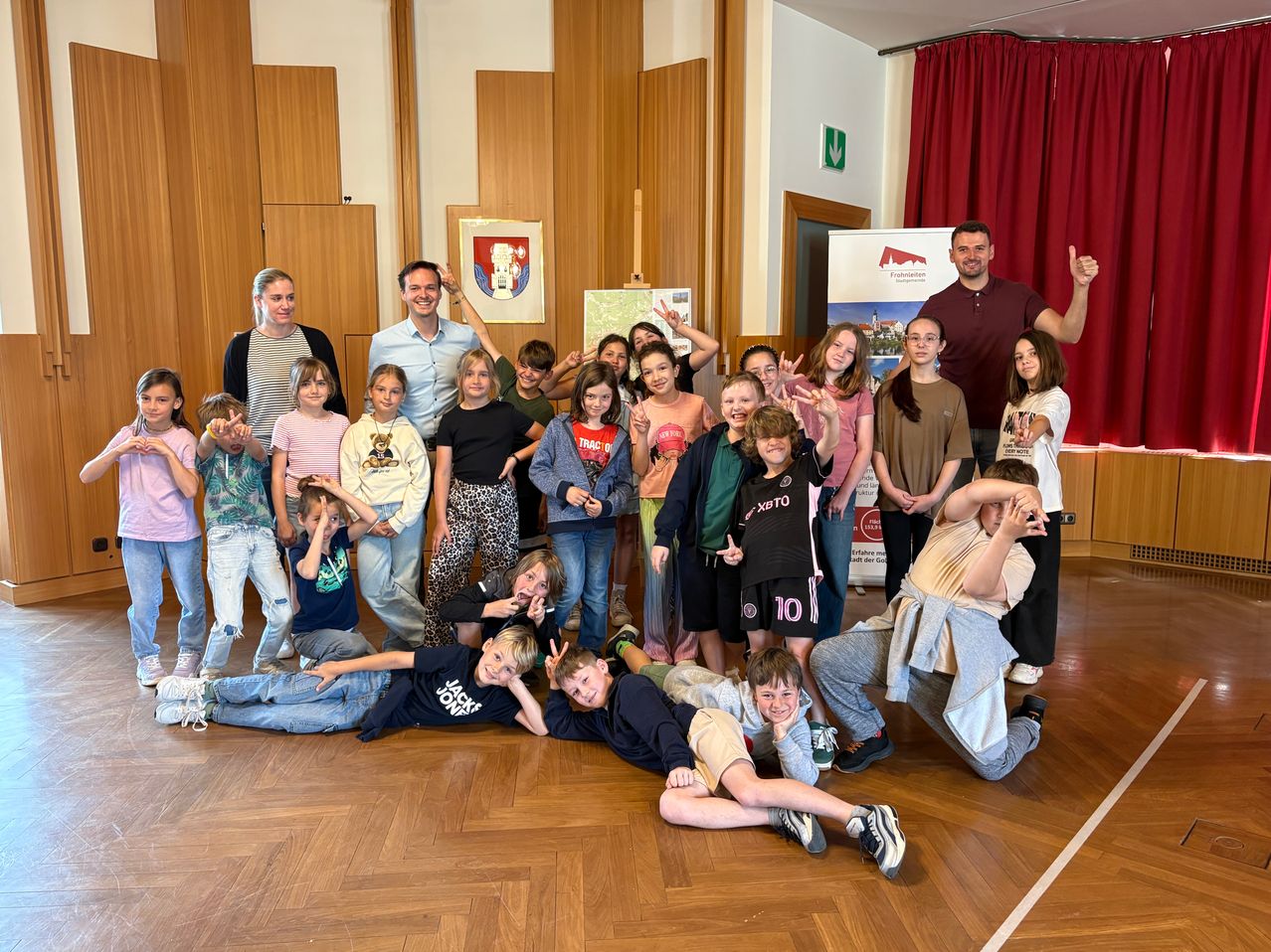 Eine Gruppe von Kindern und Erwachsenen posiert für ein Foto in einem Raum mit Holzwänden, lächelt und macht Handgesten. Ein Mann steht mit erhobener Hand.