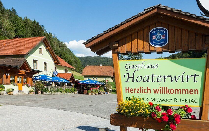 Ein einladendes Schild für Gasthaus Hoaterwirt steht draußen mit leuchtenden Blumen. Das Lokal befindet sich in einer malerischen Gegend mit Häusern und Bergen im Hintergrund.