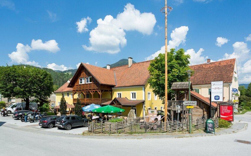 Ein gelbes Haus mit braunem Dach und Holzbalkon steht an einer Dorfkreuzung, umgeben von geparkten Autos und Grün. Ein Coca-Cola-Schild und ein Holzzaun sind in der Nähe.