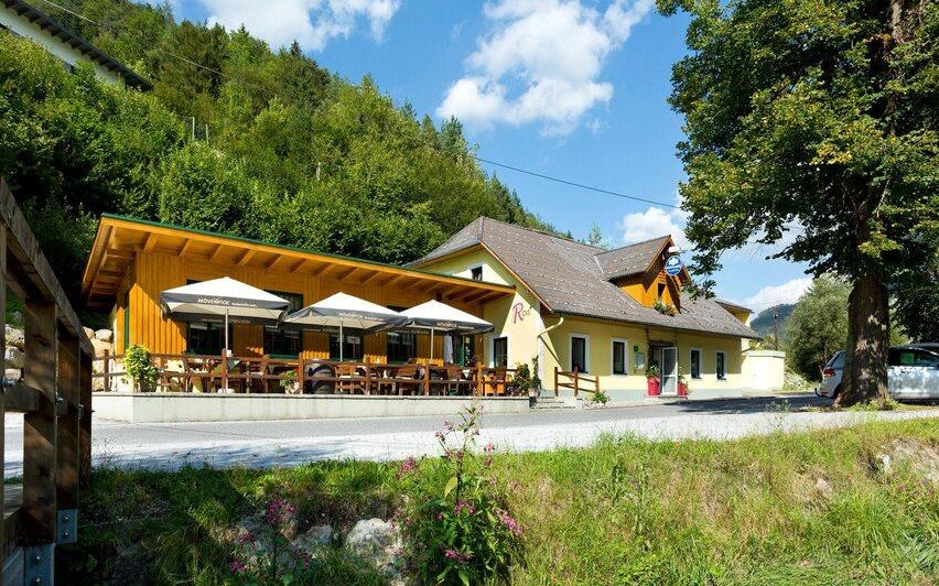 Ein gelbes Gebäude mit grauem Dach und Tischen und Schirmen draußen. Es hat ein Schild an der Wand und einen Topfpflanzen. Bäume und Berge sind im Hintergrund.