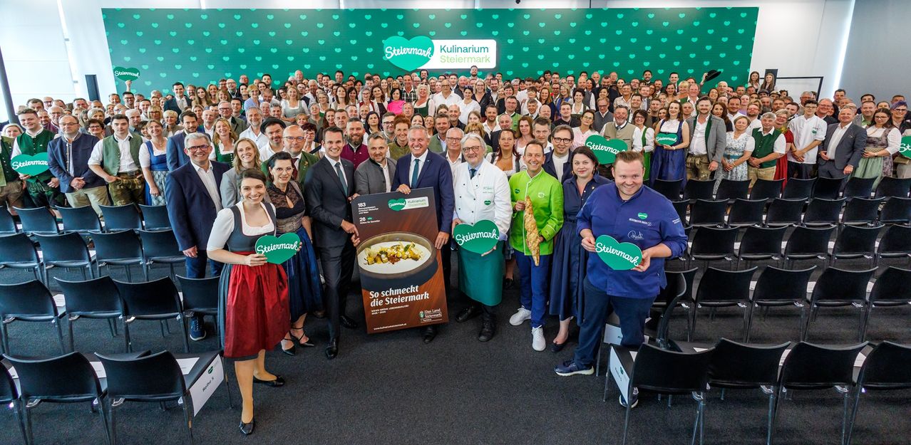 Eine Gruppe von Menschen in formeller Kleidung versammelt sich vor einem grünen Hintergrund, einige halten Schilder. Eine zentrale Figur hält ein Schild mit einem Gericht und den Worten 'So schmeckt die Steiermark'. Mehrere Köche und Personen halten grüne herzförmige Schilder.