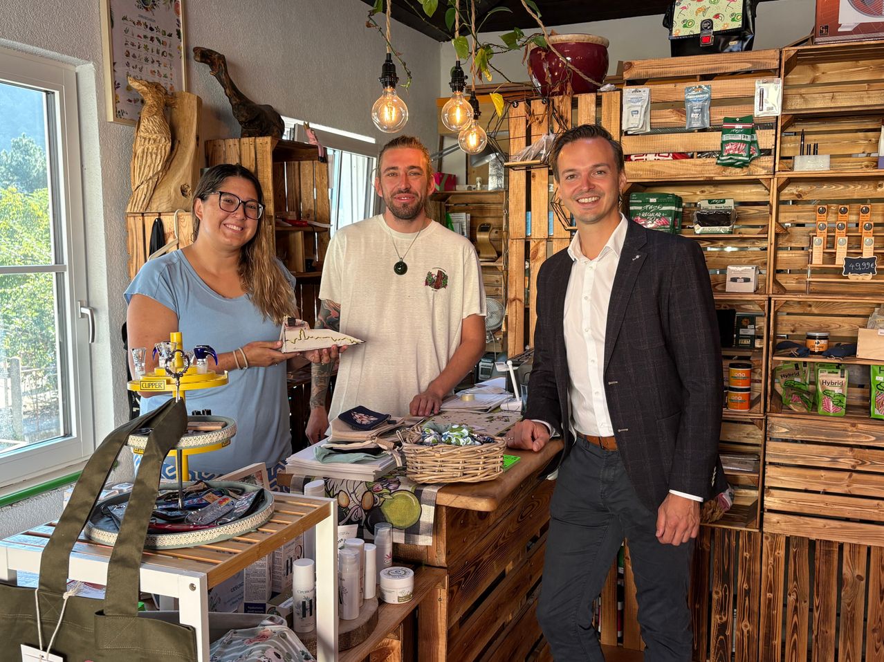 Drei Personen stehen hinter einem Holztresen in einem Geschäft. Der Mann rechts trägt Anzug und Krawatte, lächelt für das Foto. Die Frau links hält ein Stück Papier, und der Mann in der Mitte hat eine Halskette. Hinter ihnen befinden sich Regale mit verschiedenen Artikeln.