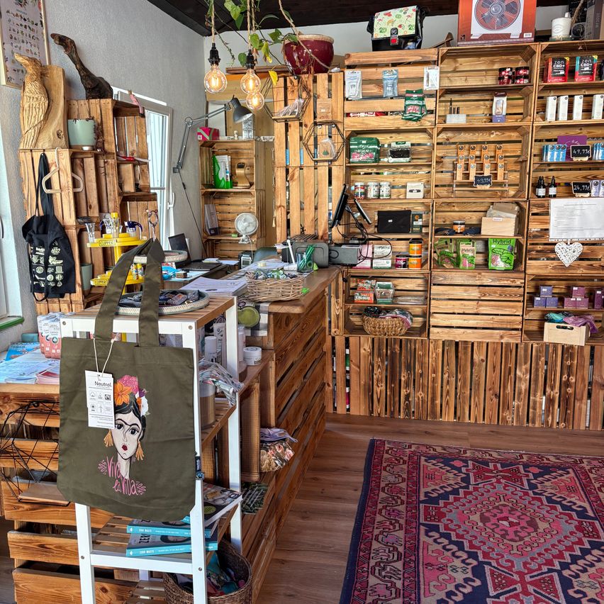 Ein Ladeninneres mit Holzregalen und Kisten, gefüllt mit verschiedenen Produkten. Eine grüne Tragetasche mit Frida-Kahlo-Design hängt an einem weißen Ständer. Ein Teppich liegt auf dem Boden.