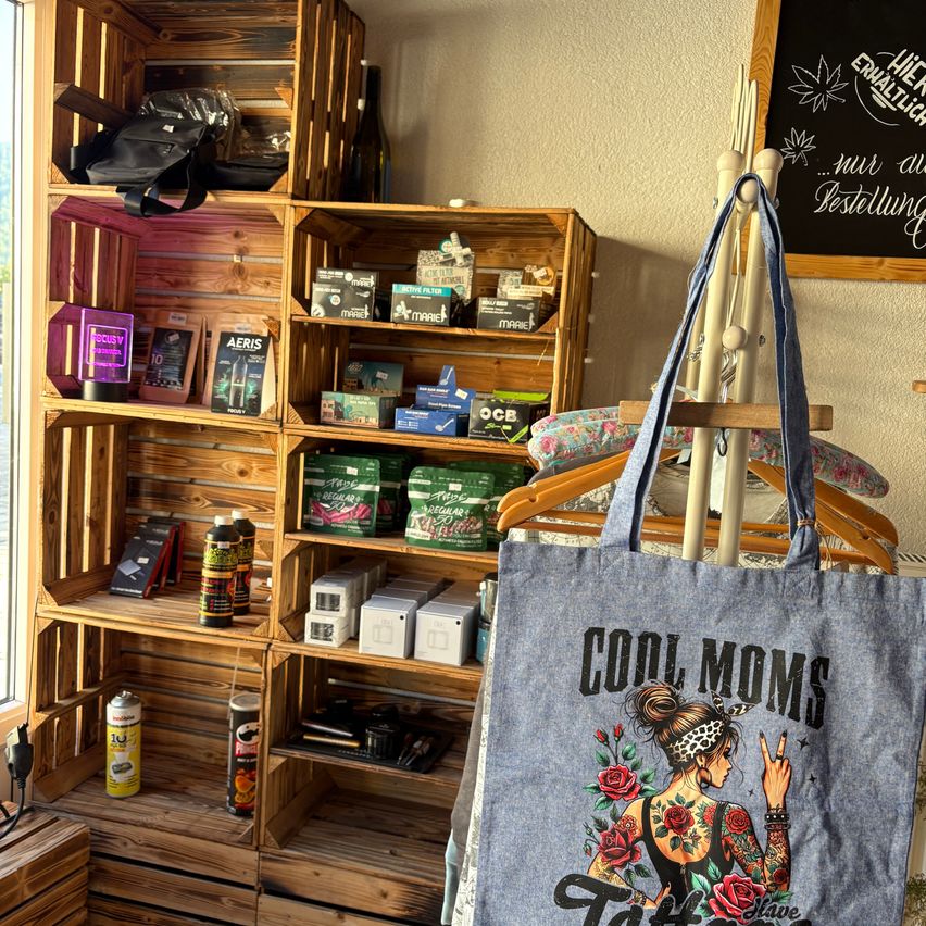 Ein Raum mit Holzregalen zeigt verschiedene Produkte, darunter Flaschen, Schachteln und Taschen. Ein Schild an der Wand mit der Aufschrift 'Hier enthaltlich... nur auf Bestellung'. Eine Jeanstasche mit dem Text 'COOL MOMS' hängt an einem Kleiderbügel.