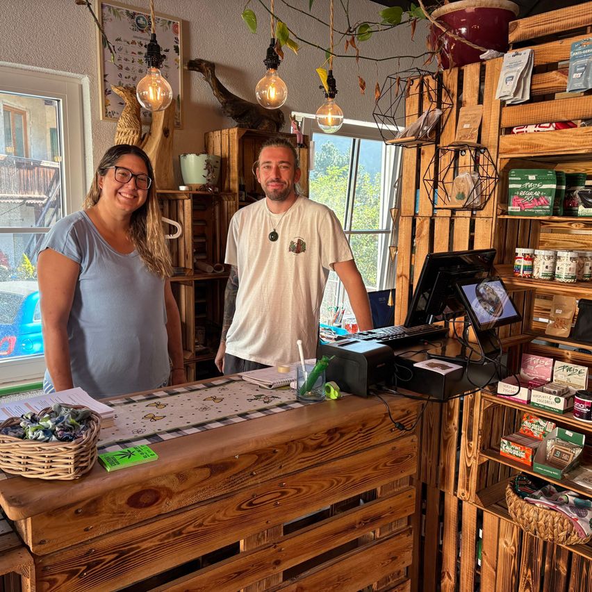 Ein Mann und eine Frau stehen hinter einer Theke in einem Geschäft mit Holzregalen. Die Theke hat einen Monitor, eine Tastatur und verschiedene Gegenstände. Ein Korb mit Süßigkeiten steht auf der Theke.