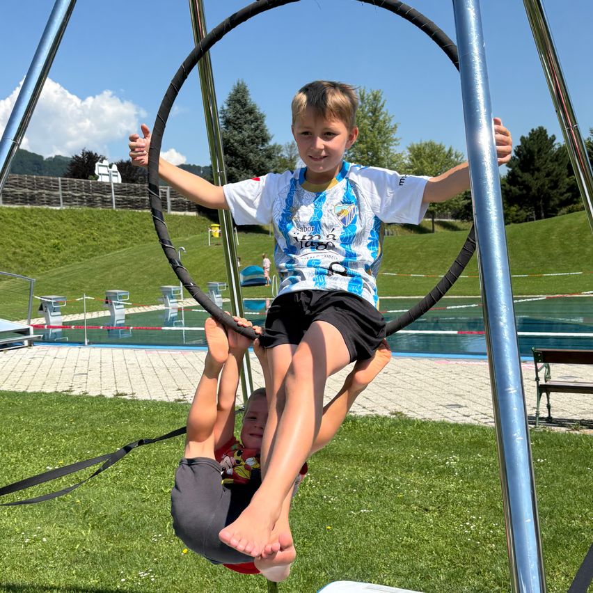 Ein junger Junge hält ein jüngeres Kind in einer Schaukel auf einem Außengelände mit einem Schwimmbecken, Bäumen und Bergen im Hintergrund.