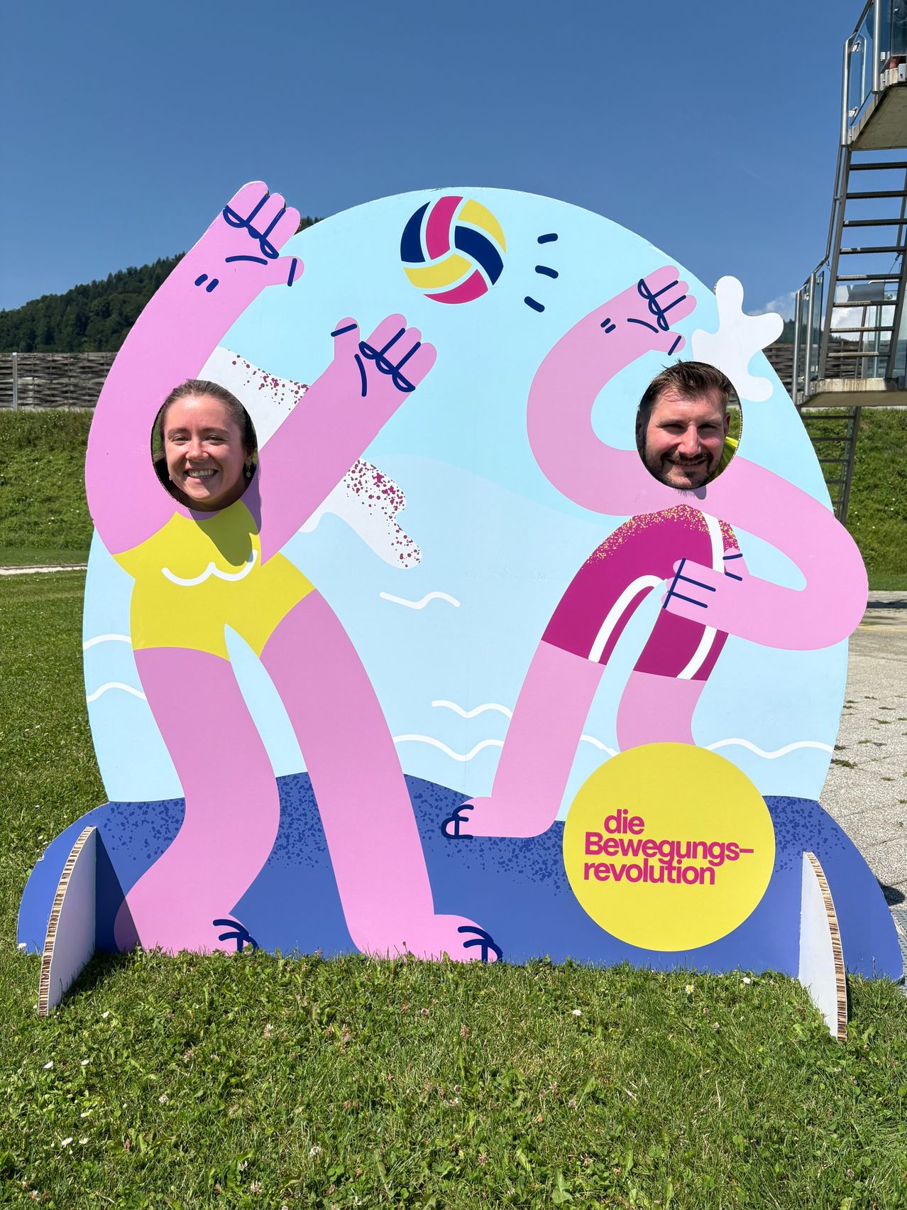 Ein farbenfroher Ausschnitt-Fotostand zeigt zwei Menschen, die Volleyball spielen, mit dem Text 'die Bewegungs-revolution' in einem gelben Kreis.