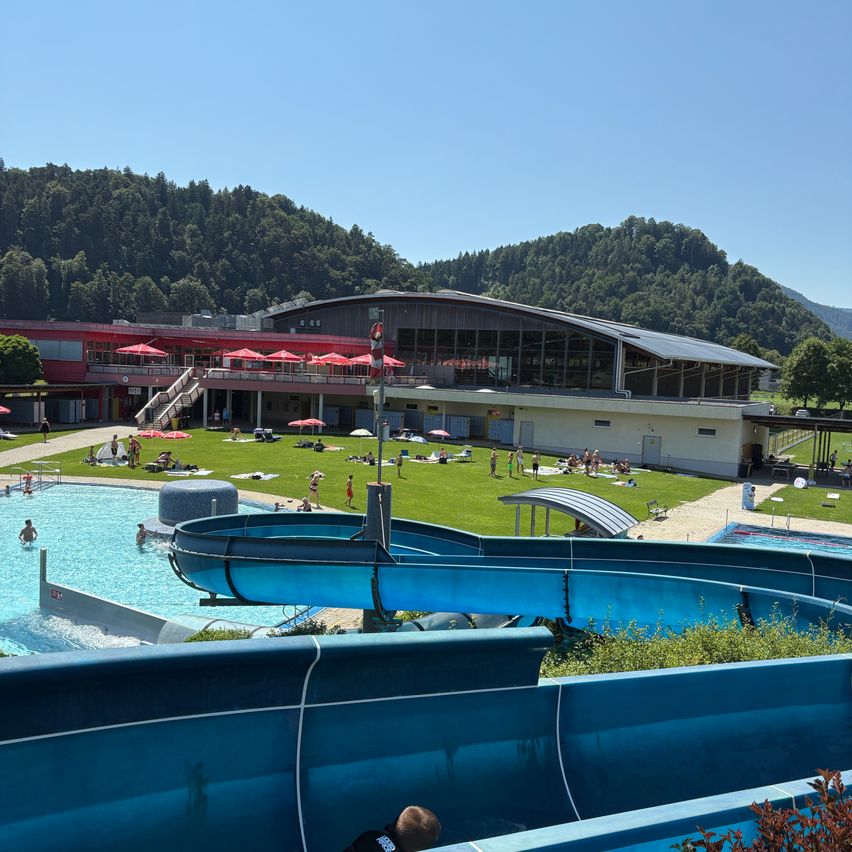 Ein Freibad mit einem großen Pool und blauen Rutschen. Menschen entspannen auf der Wiese und unter Sonnenschirmen. Ein großes Gebäude mit vielen Fenstern ist im Hintergrund.