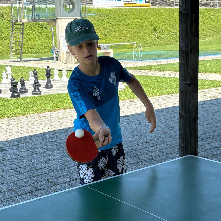 Ein Junge spielt im Freien Tischtennis, steht vor einem Tisch mit einem roten Schläger. Ein Schachset ist auf dem Rasen hinter ihm platziert.