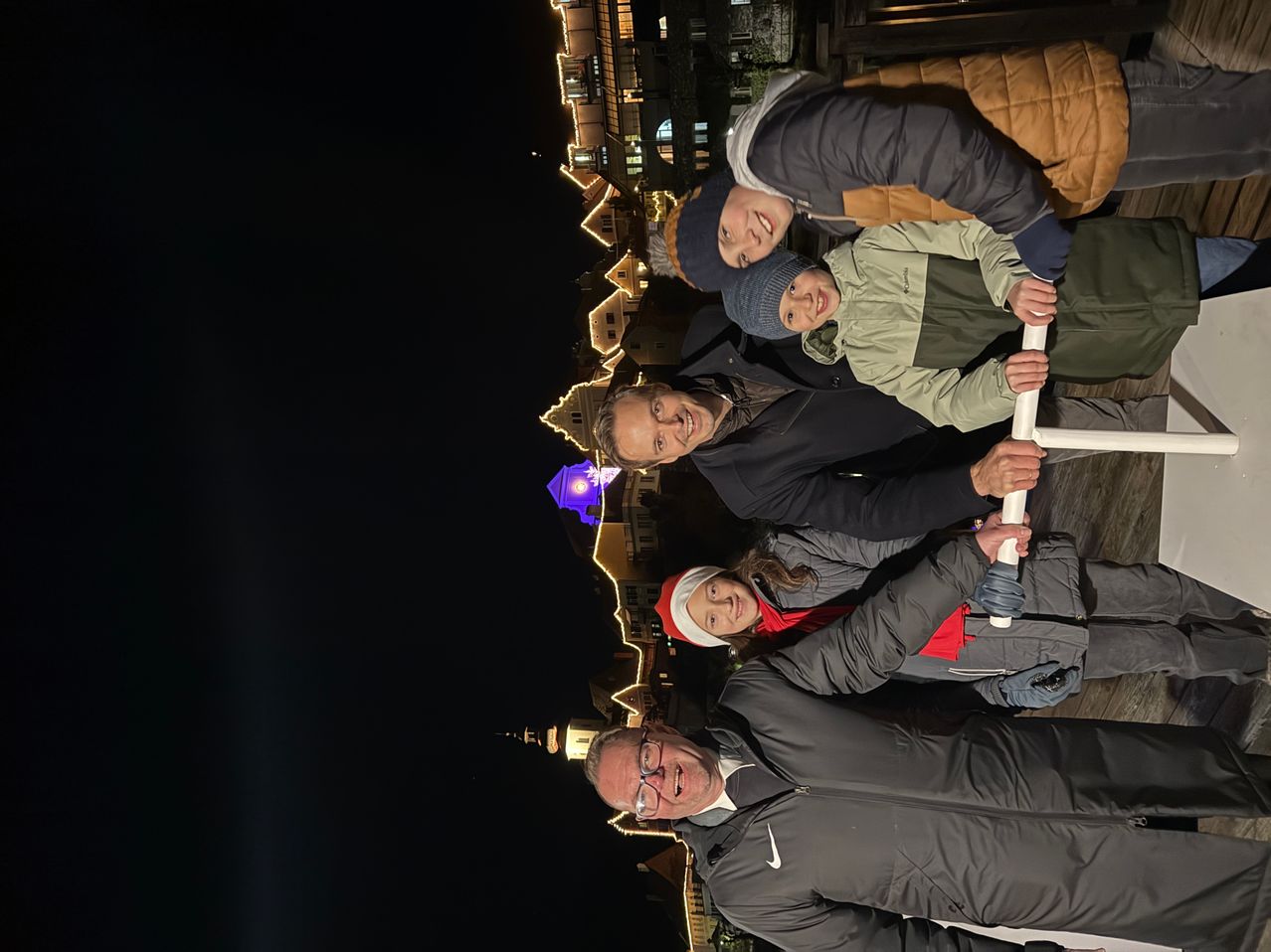 Eine Gruppe von Menschen, einschließlich Erwachsener und Kindern, steht bei Nacht auf einer Brücke. Sie lächeln und scheinen für ein Foto zu posieren. Hinter ihnen sind Gebäude mit Lichtern.