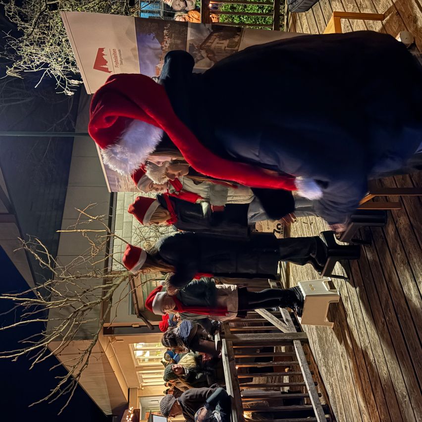 Eine Gruppe von Menschen in Weihnachtsmützen versammelt sich auf einer Holzterrasse, einige stehend und andere auf Treppenstufen sitzend. Eine Person lehnt sich an ein Geländer.
